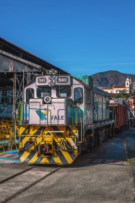 A vintage diesel train with the number 641 and a white, green, and yellow livery is stationed on railway tracks outdoors during daylight hours, with a clear blue sky overhead. The front of the train shows visible headlights, control windows, and safety railings, with the word 'VALE' displayed on the lower front section. Behind the train, a few freight containers are loaded onto the carriages, and in the background, there are low-rise buildings, a church with a steeple, and mountains in the distance. This scene suggests a transport or logistics environment relevant to moving and relocation services, with the train potentially used for furniture transport or equipment transit as part of home relocation processes. The lighting highlights the metallic surfaces and the various textures of the train and surrounding infrastructure, reflecting typical railway yard conditions. Maida Vale Man and Van's services may include coordination with such transport modes for efficient house removals.