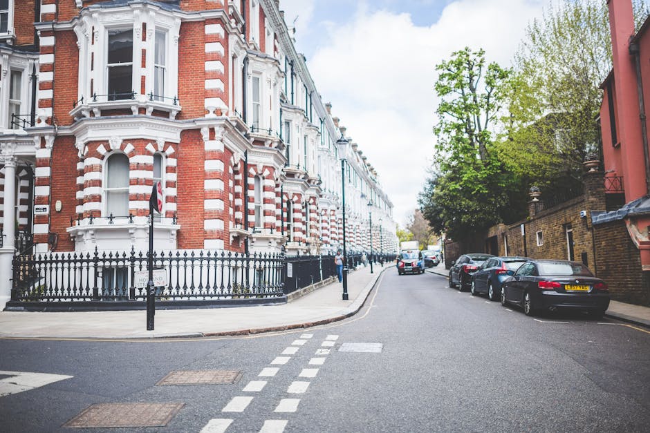 A quiet residential street scene in Maida Vale featuring a row of Victorian-style terraced houses with red brick facades, white decorative trim, and bay windows. The buildings are positioned alongside the pavement, which is accessible for moving crews. Parked cars line the street on the right side, including a black convertible, a silver sedan, and other vehicles, indicating available parking for house removals. On the left, there is a black iron fence surrounding a corner property with a sign indicating no parking during certain hours. The street is narrow, with dashed white lines marking the vehicle lane, and is partially shaded by trees with green foliage. Clear blue sky is visible above, and a few pedestrians are present in the distance. The scene captures an outdoor environment typical for home relocation logistics, with space for loading and unloading furniture and packed boxes, showcasing the type of setting Maida Vale Man and Van services operate within during furniture transport and packing and moving procedures.