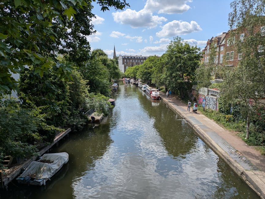 A daytime view of the Little Venice canal lined with leafy green trees on both sides. The water reflects the blue sky and white clouds above. Parked boats are visible along the canal's edge, with one covered in a protective tarp. A paved pathway runs parallel to the water on the right side, where a few pedestrians are walking. On the right, there are brick residential buildings with large windows, some partially obscured by the trees. In the background, a church steeple and additional buildings can be seen, indicating an urban setting. This scene exemplifies a peaceful canal-side environment typical of Maida Vale, often associated with house removals or furniture transport in the area, as supported by Maida Vale Man and Van's services connected to house relocations and moving logistics.