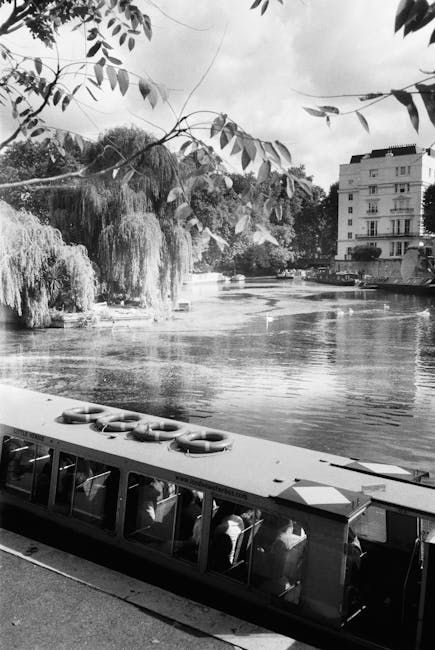 A daytime view of the Little Venice canal lined with leafy green trees on both sides. The water reflects the blue sky and white clouds above. Parked boats are visible along the canal's edge, with one covered in a protective tarp. A paved pathway runs parallel to the water on the right side, where a few pedestrians are walking. On the right, there are brick residential buildings with large windows, some partially obscured by the trees. In the background, a church steeple and additional buildings can be seen, indicating an urban setting. This scene exemplifies a peaceful canal-side environment typical of Maida Vale, often associated with house removals or furniture transport in the area, as supported by Maida Vale Man and Van's services connected to house relocations and moving logistics.