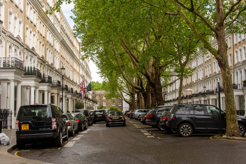 Image showing a street scene in Maida Vale with residential buildings on both sides and a historic clock tower in the background under a clear blue sky. On the left side, there is a sidewalk with a bl