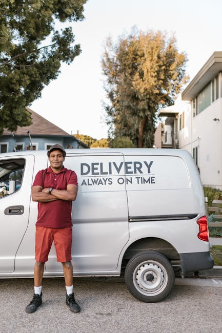 A man wearing a white t-shirt, dark pants, and a baseball cap is walking next to a white commercial van parked on the side of a residential street. The van, associated with Maida Vale Man and Van's re